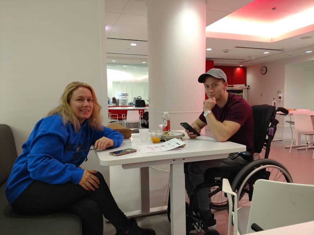 Cosmo and his Mom sitting at a table together. Cosmo is in a wheelchair and has his phone in hand.